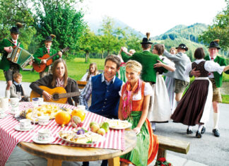 Ein Paar sitzt an einem reich gedeckten Tisch im Freien, umringt von Musikern und Tänzern (Symbolbild zum Salzburger Bauernherbst)