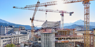 Blick auf das Baustellenareal DAS RAIQA vom Norden aus mit Dachlandschaft Bauteil B und dem unterstellen Schalungstisch für Hammerkopf im linken Vordergrund. Im Bild rechts der ebenfalls mit Gerüsttürmen unterstellte Eingangsbereich des neuen hybrid genutzten Haupthauses sowie im Hintergrund die Stahlbeton-Skelett-Struktur des alten RLB-Turms in Revitalisierung.