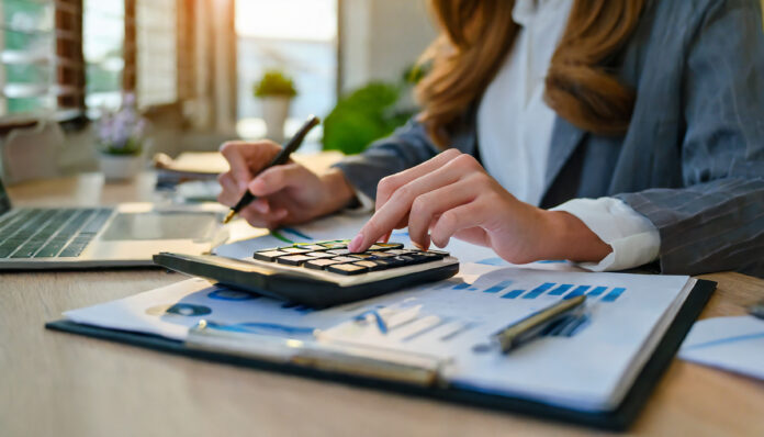 Weibliche Hände an einem Tisch mit Taschenrechner, Zettel und Stift als Symbolbild für Finanzbildung