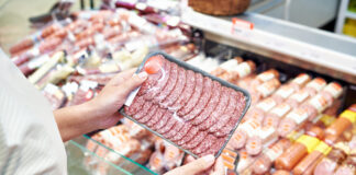 Eine Person hält abgepackten Wurstaufschnitt vor einer Wursttheke, als Symbolbild für Wurstwaren