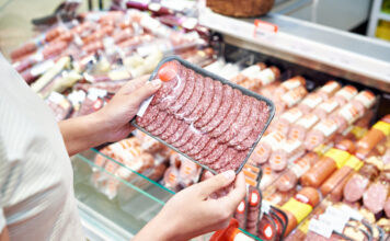 Eine Person hält abgepackten Wurstaufschnitt vor einer Wursttheke, als Symbolbild für Wurstwaren