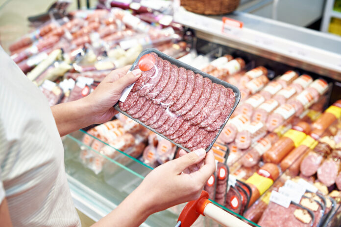 Eine Person hält abgepackten Wurstaufschnitt vor einer Wursttheke, als Symbolbild für Wurstwaren