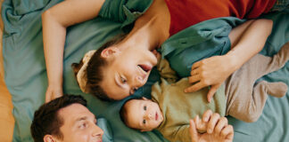 Eine Familie – Mutter, Vater und Baby – liegen auf einem Bett, als Symbolbild für Kindervorsorge