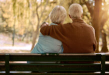 Love, hug and old couple in a park on a bench for a calm, peaceful or romantic summer marriage anniversary date. Nature, romance or back view of old woman and elderly partner in a relaxing embrace.