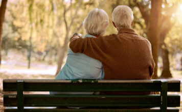 Love, hug and old couple in a park on a bench for a calm, peaceful or romantic summer marriage anniversary date. Nature, romance or back view of old woman and elderly partner in a relaxing embrace.