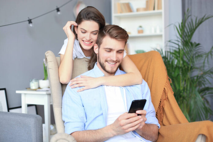 Junges Paar beim Online-Shopping zu Hause, mit dem Smartphone auf dem Sofa.