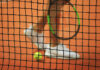 Tennisnetz im Fokus, unscharfer Spieler und Ball im Hintergrund.