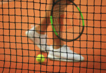 Tennisnetz im Fokus, unscharfer Spieler und Ball im Hintergrund.