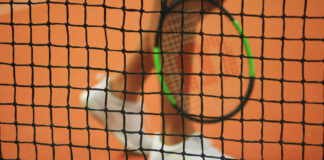 Tennisnetz im Fokus, unscharfer Spieler und Ball im Hintergrund.