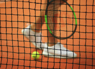 Tennisnetz im Fokus, unscharfer Spieler und Ball im Hintergrund.