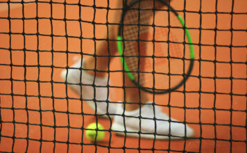 Tennisnetz im Fokus, unscharfer Spieler und Ball im Hintergrund.