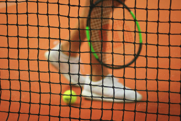Tennisnetz im Fokus, unscharfer Spieler und Ball im Hintergrund.