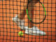 Tennisnetz im Fokus, unscharfer Spieler und Ball im Hintergrund.