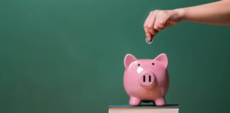 Person depositing money in a pink piggy bank on top of books with chalkboard in the background as concept image of the costs of education