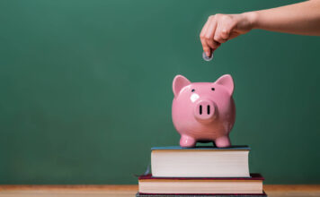 Person depositing money in a pink piggy bank on top of books with chalkboard in the background as concept image of the costs of education