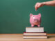 Person depositing money in a pink piggy bank on top of books with chalkboard in the background as concept image of the costs of education