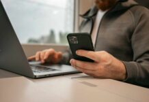 Ein Mann sitzt vor einem Laptop mit seinem Smartphone in der Hand, als Symbolbild für Online Banking