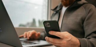 Ein Mann sitzt vor einem Laptop mit seinem Smartphone in der Hand, als Symbolbild für Online Banking
