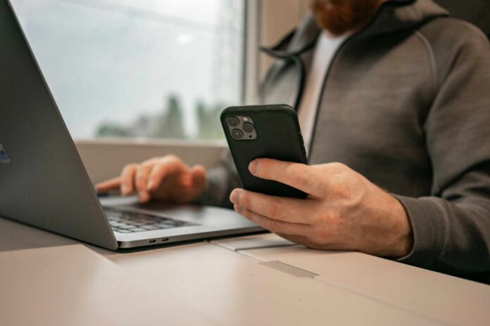 Ein Mann sitzt vor einem Laptop mit seinem Smartphone in der Hand, als Symbolbild für Online Banking