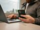 Ein Mann sitzt vor einem Laptop mit seinem Smartphone in der Hand, als Symbolbild für Online Banking