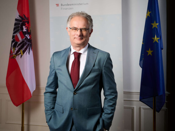 Finanzminister Markus Marterbauer stehend zwischen einer Österreich- und EU-Flagge