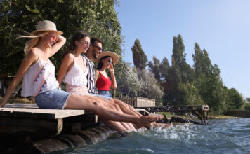 Sommer genießen statt warten müssen Drei Frauen und ein Mann sitzen auf einem Steg und lassen die Beine ins Wasser hängen