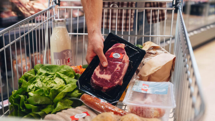 Jemand legt ein abgepacktes Stück Fleisch in einen halbvollen Einkaufswagen.